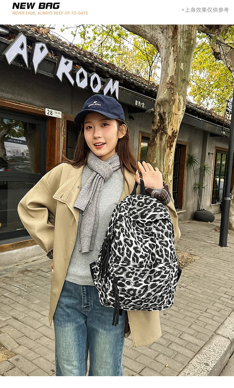 A woman wearing a beige coat and jeans holds a black leopard print canvas backpack outdoors.