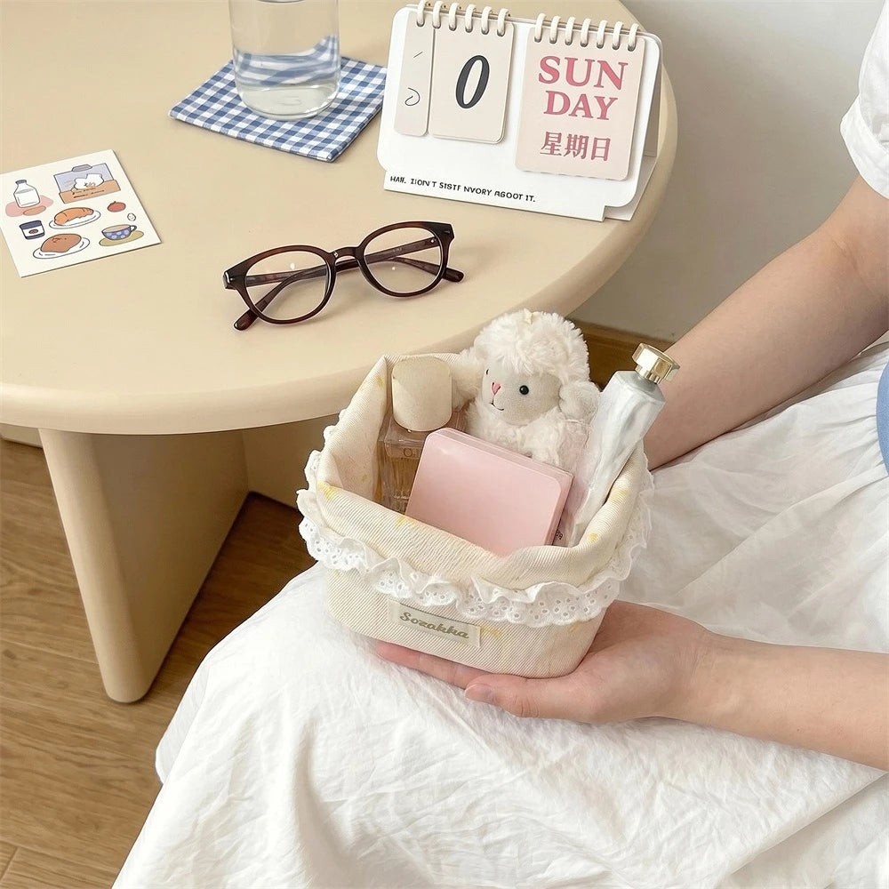 A light pink fabric storage basket with a heart and bow is shown next to a cup of water and stationery on a table.