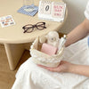 A light pink fabric storage basket with a heart and bow is shown next to a cup of water and stationery on a table.