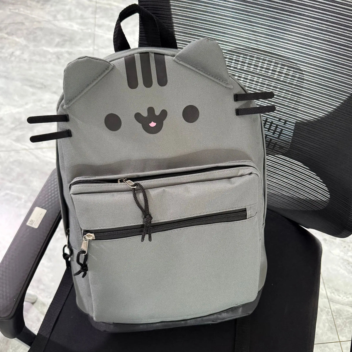 A gray Pusheen cat canvas backpack with black whiskers and eyes resting on a black chair.