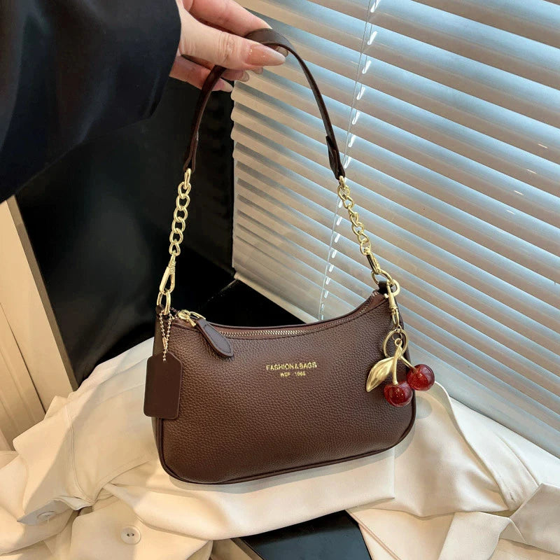 A brown leather crescent crossbody bag with a gold chain strap is displayed against a white shirt.