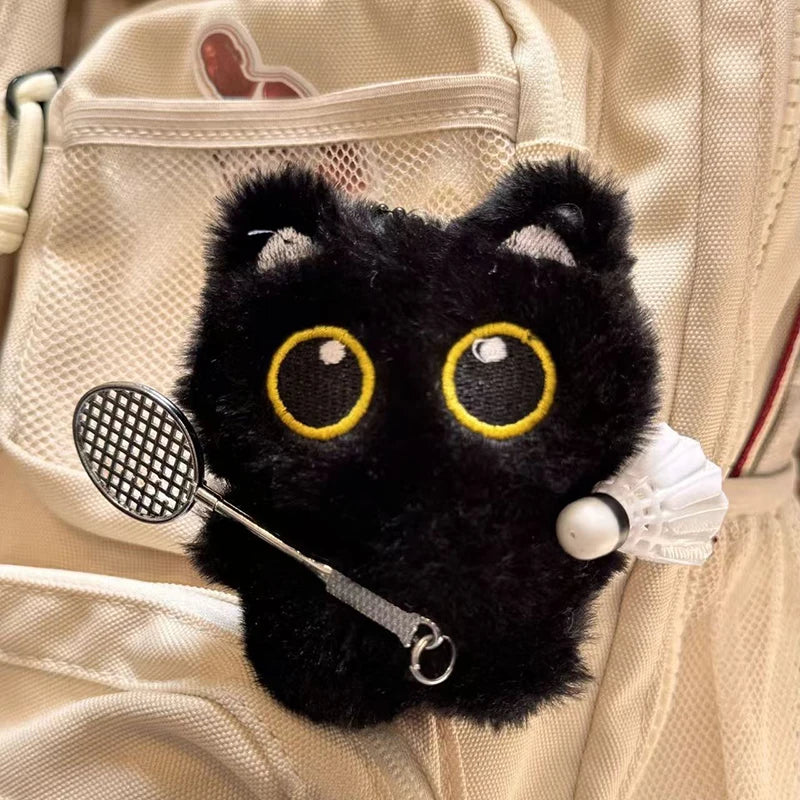Colgante de gato negro de peluche con ojos amarillos grandes sujeto a una bolsa de tela blanca.