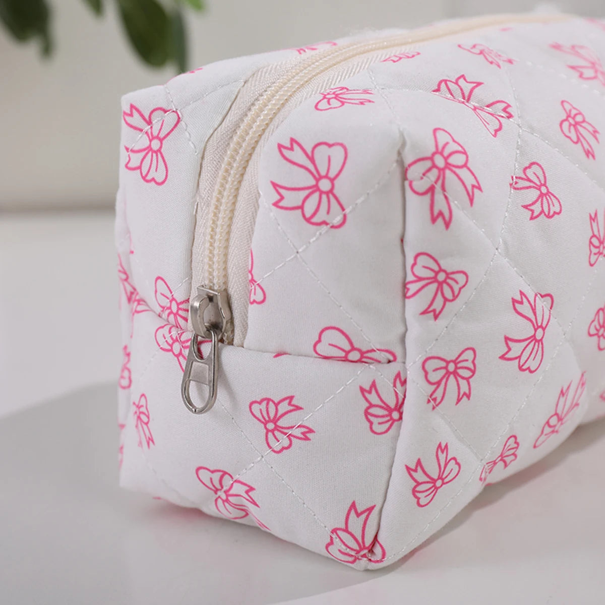 Bolsa acolchada blanca para maquillaje con estampado de lazo rosa y cremallera plateada.