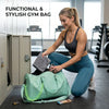 A woman is sitting on the floor with a light green gym bag and a dark gray towel in a gym setting.
