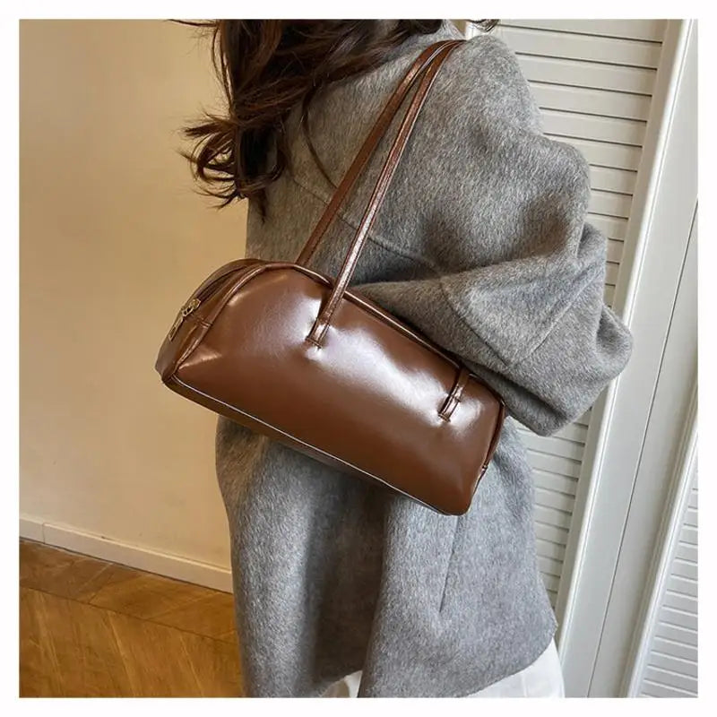 A brown glossy PU leather tote bag with dual straps is shown carried in front of a person wearing a gray coat.