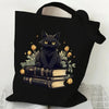 Black canvas tote bag featuring a black cat sitting on a stack of old books with golden leaves and flowers.