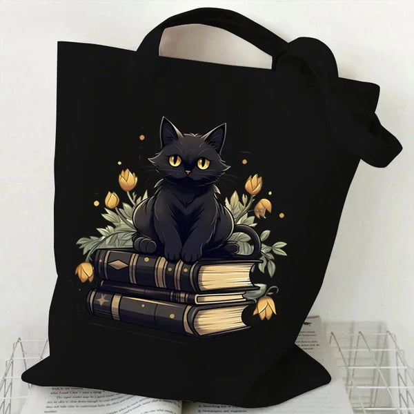 Black canvas tote bag featuring a black cat sitting on a stack of old books with golden leaves and flowers.