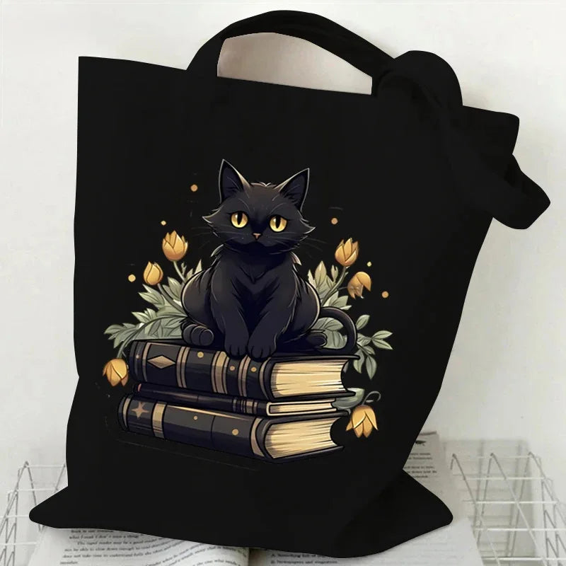 Black canvas tote bag featuring a black cat sitting on a stack of old books with golden leaves and flowers.