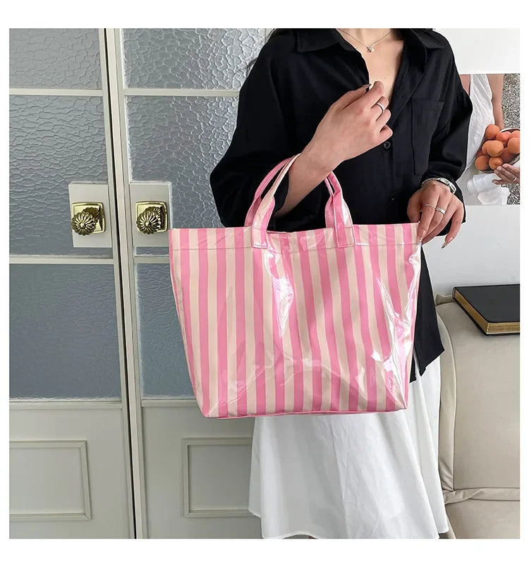 A pink and white striped waterproof PVC shoulder tote bag is held by a person in front of a glass door.