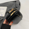 A hand holds a black vegan PU crossbody bag with gold hardware against a gray fabric background.