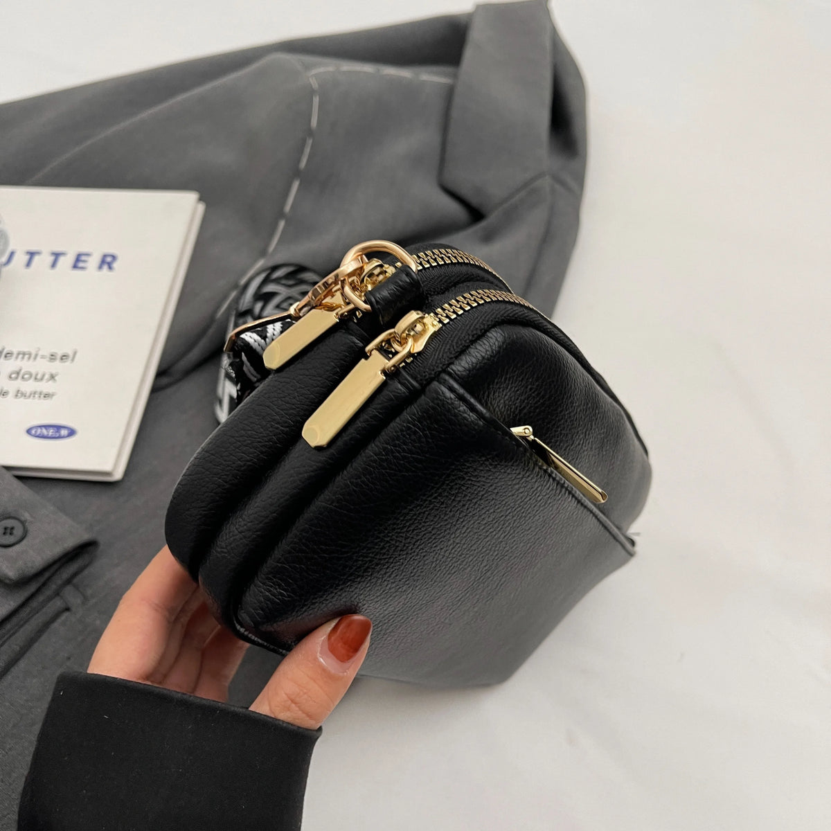 A hand holds a black vegan PU crossbody bag with gold hardware against a gray fabric background.