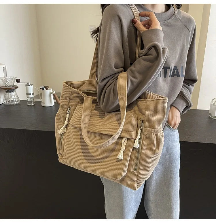 A woman is holding a beige corduroy shoulder bag with a zipper on her shoulder while standing in front of a dark counter.