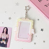 A clear display case framed in pink and yellow, featuring angel wings, suspended by a silver charm.