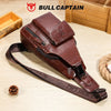 A brown genuine leather sling backpack with a dark brown strap is displayed on a wooden surface.