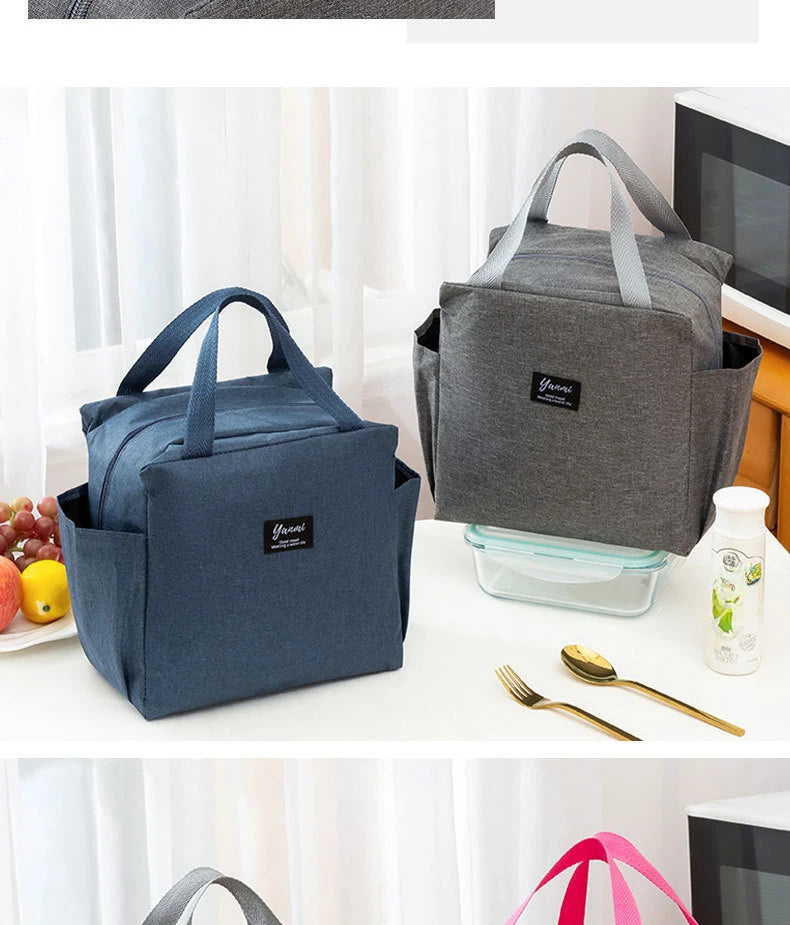 A dark blue insulated canvas tote bag with double pockets resting on a white surface next to a laptop and a glass container.