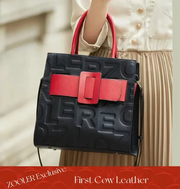 A black leather shoulder bag with a red accent strap and gold hardware being held by a person.