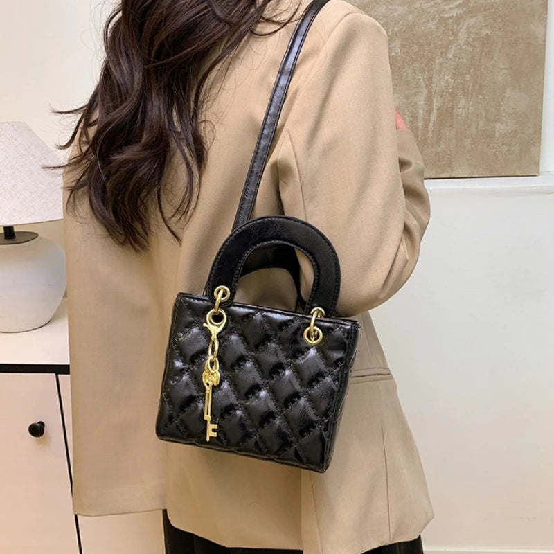 A black quilted crossbody handbag with gold chain accents carried by a person wearing a beige blazer.