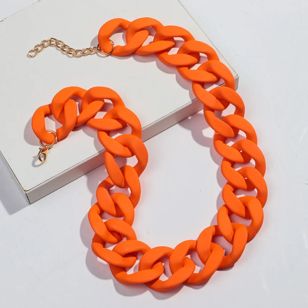 An orange matte acrylic choker necklace with a gold-colored chain is displayed on a white box.