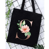 Black canvas tote bag featuring a large gold script letter S surrounded by pink floral greenery