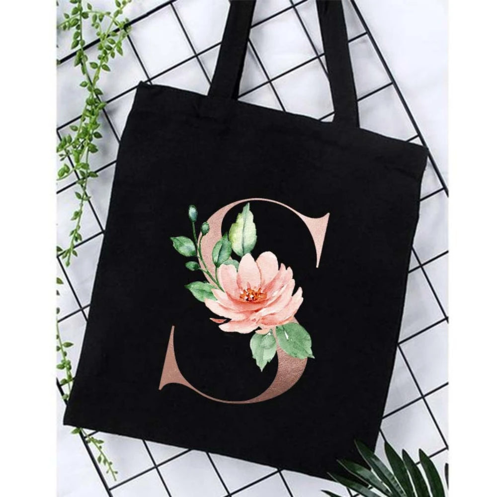 Black canvas tote bag featuring a large gold script letter S surrounded by pink floral greenery