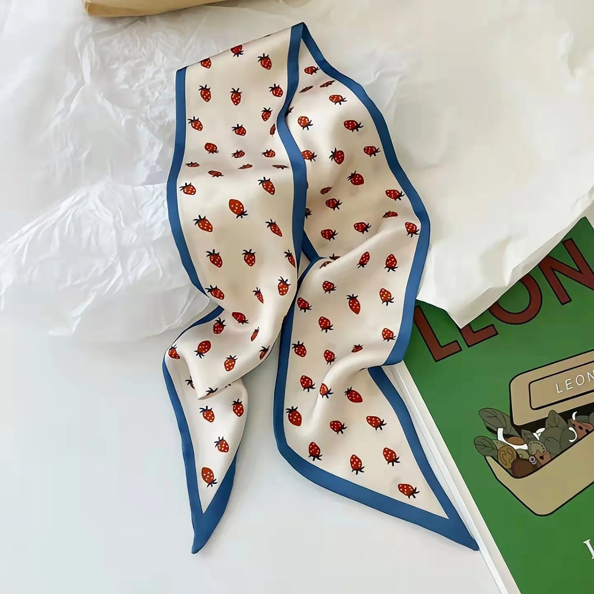 Écharpe de soie couleur crème avec bordure bleue et motif fraise rouge, présentée sur du papier blanc.