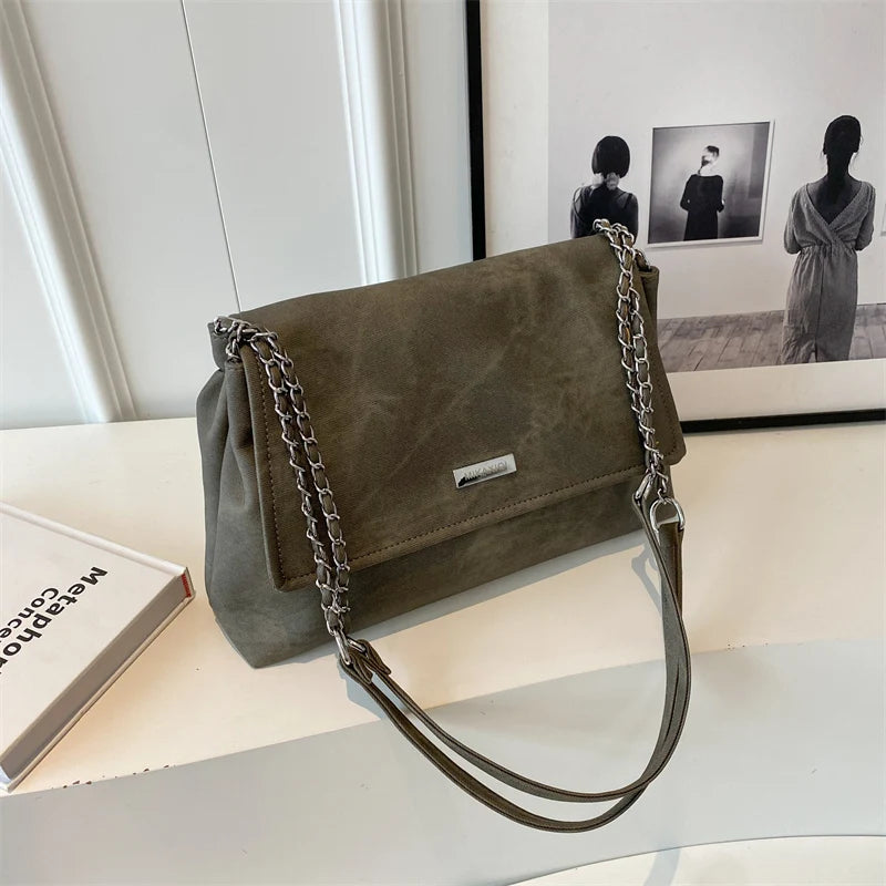 Bolso de cuero retro verde oliva oscuro con cadena colgado sobre una superficie blanca junto a una foto enmarcada.