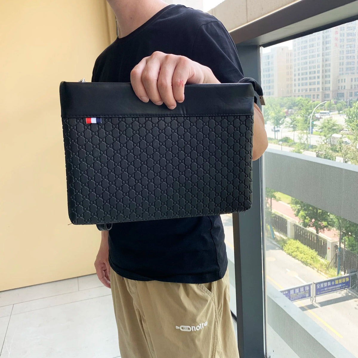 A person holds a black PU leather clutch bag with a flap and a small flag detail against a light wall.