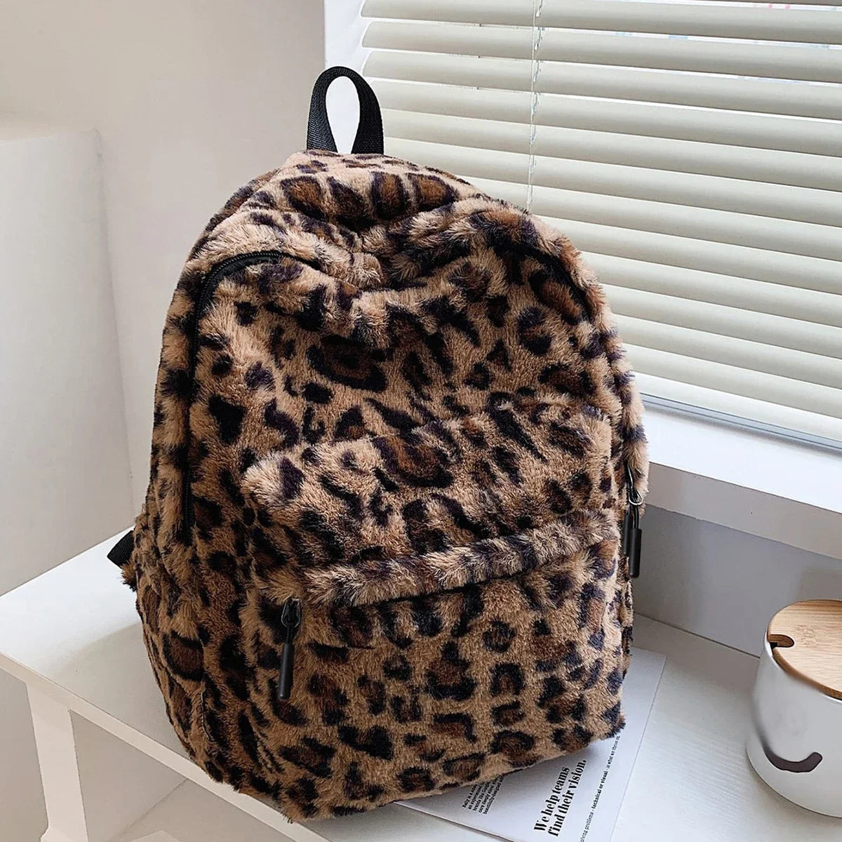 A brown leopard print plush backpack with black straps resting on a white surface next to a window blind.