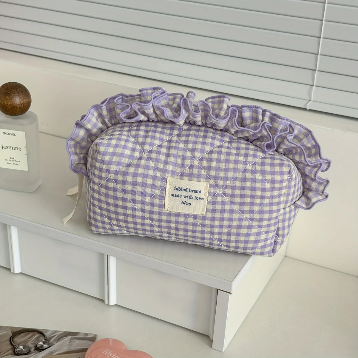 A lavender checkered makeup pouch with a ruffled lace trim resting on a white shelf.