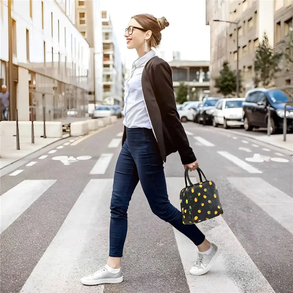 Une femme en blazer foncé et jeans marche sur un passage piéton tenant un sac à lunch en tissu noir.