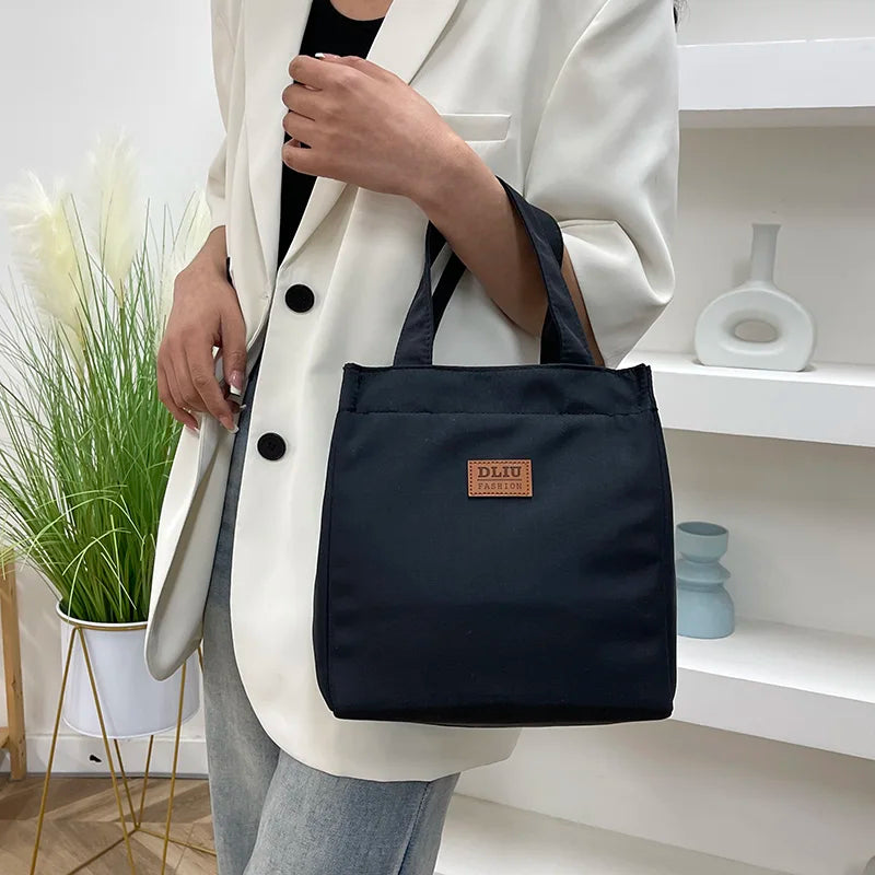 A woman holds a black insulated lunch bag in front of a white blazer and light blue jeans.