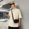 A young man wearing a beige t-shirt and a yellow cap holds a black smartphone while leaning against a gray wall.