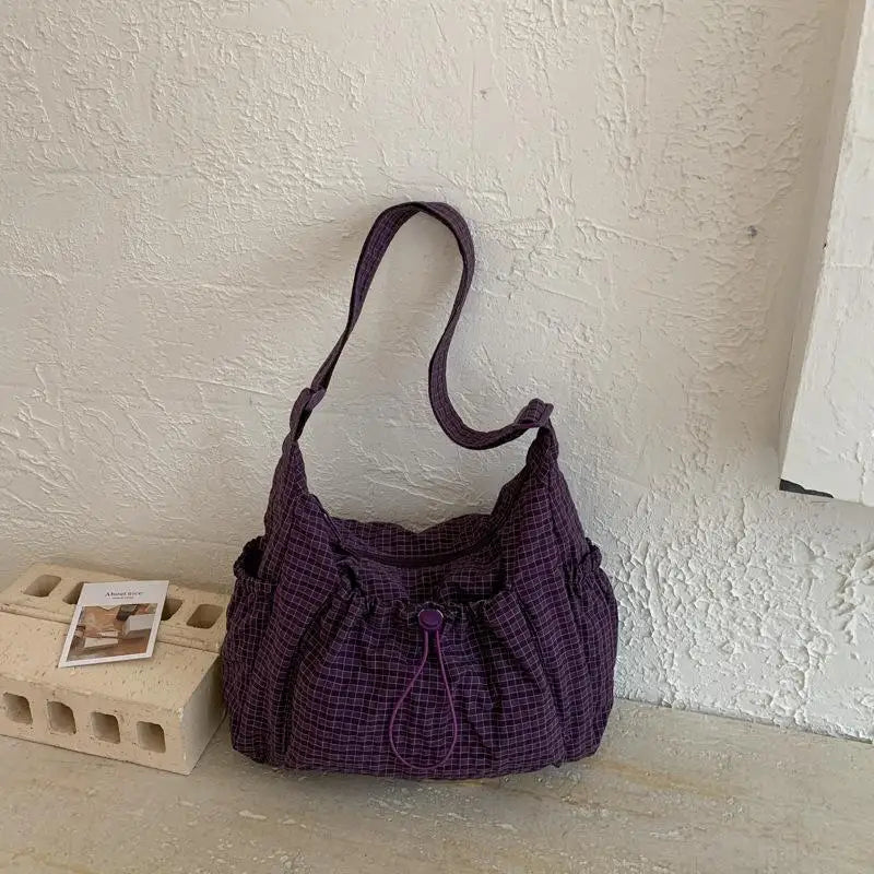 A purple plaid nylon drawstring tote bag is displayed on a light surface against a white wall.