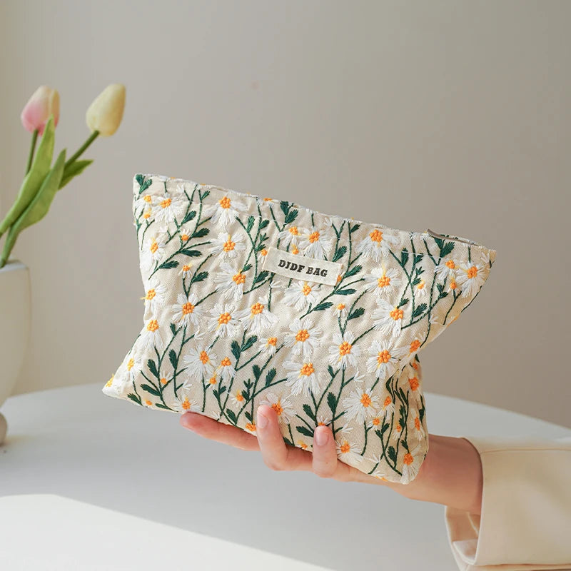 A hand holds a floral canvas makeup bag featuring green and orange flowers against a light background.