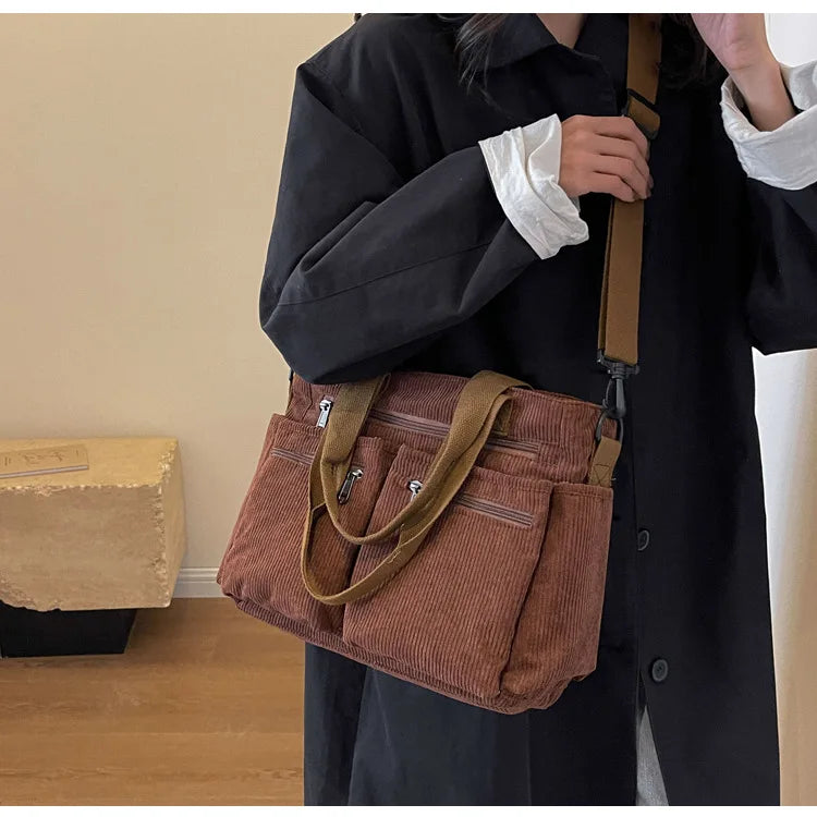 A person is holding a brown textured corduroy tote bag with adjustable straps against a black coat.
