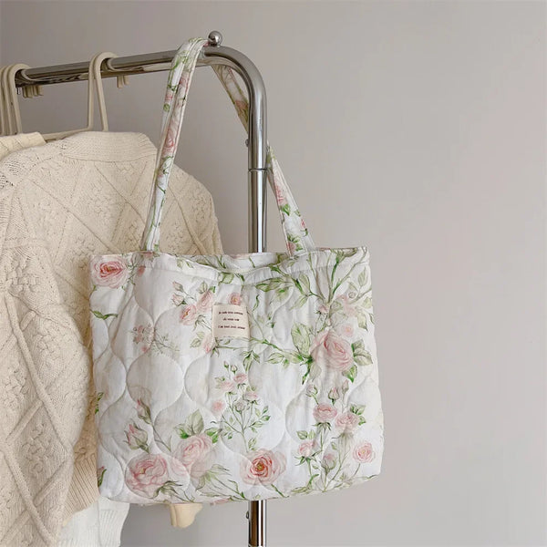 Bolso de tela acolchada blanca con bordado floral rosa y verde, colgado en un soporte de metal.