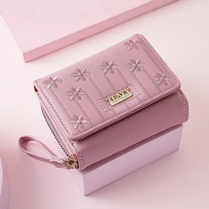 A pink PU leather compact wallet with a zipper and decorative floral stitching resting on a light pink surface.
