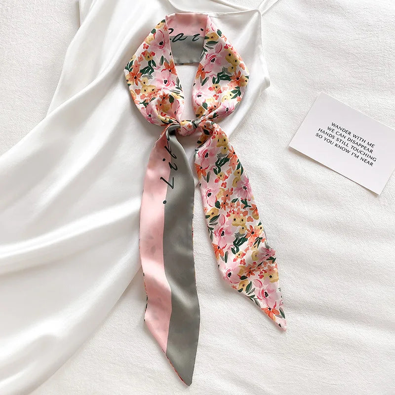 Écharpe florale de soie rose et vert nouée en nœud, posée sur un tissu blanc avec une petite carte.
