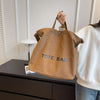 A person holds a brown canvas tote bag with blue lettering on a dark surface.
