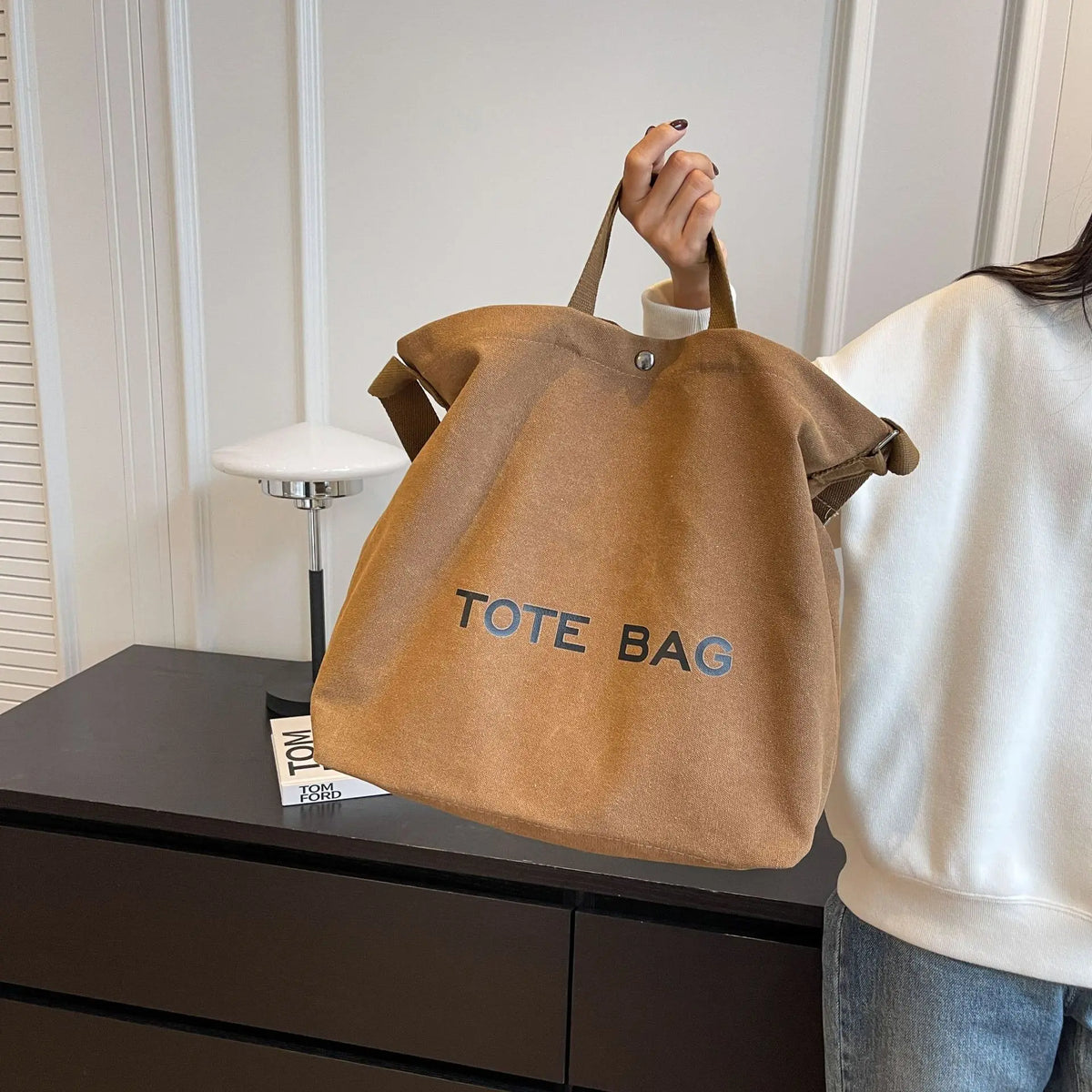 A person holds a brown canvas tote bag with blue lettering on a dark surface.