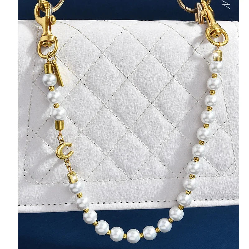 A pearl and gold letter charm necklace displayed against a white quilted bag.