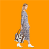 A woman wearing a blue floral dress and white boots holds a brown and white cow print leather tote bag against an orange back