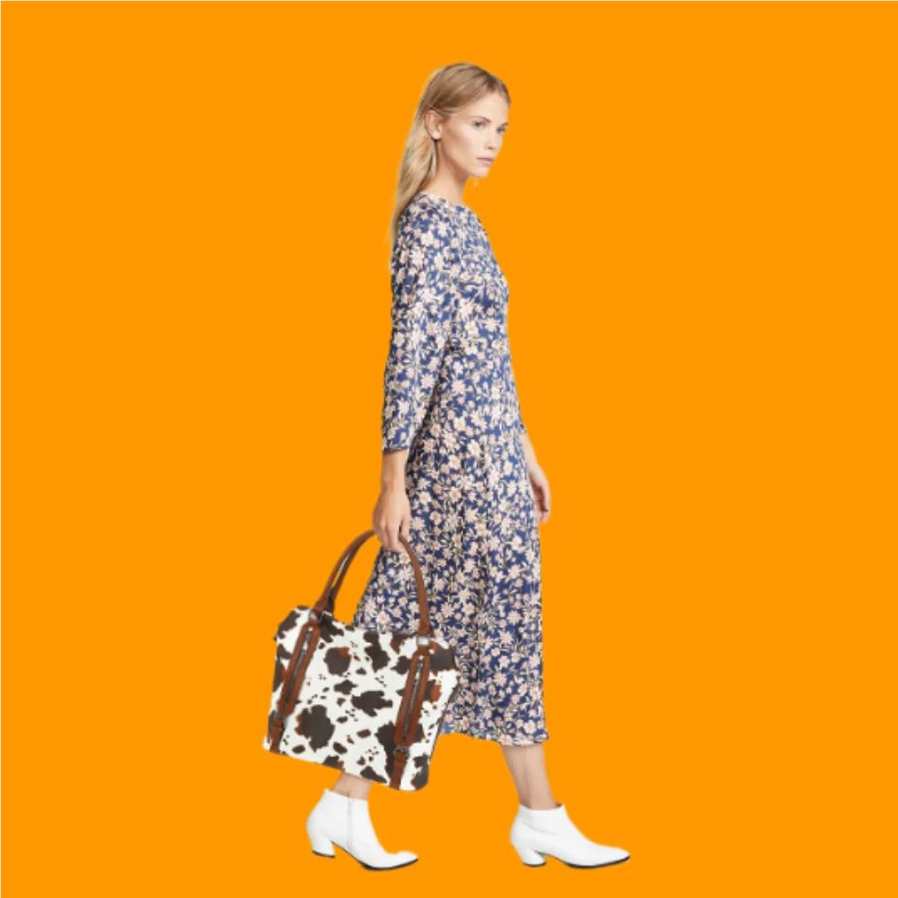 A woman wearing a blue floral dress and white boots holds a brown and white cow print leather tote bag against an orange back
