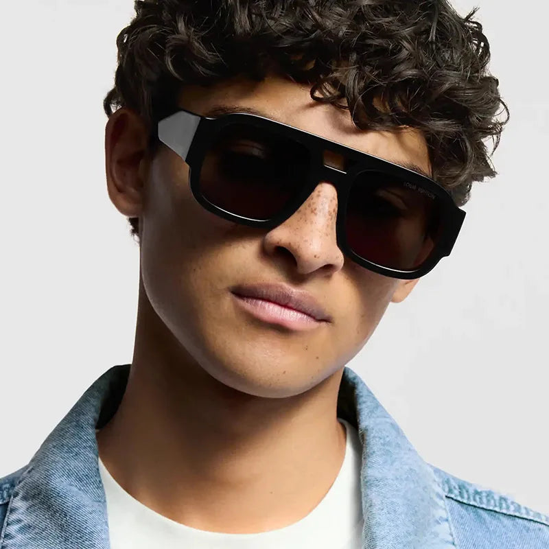 A male model wearing black oversized square sunglasses and a denim jacket.