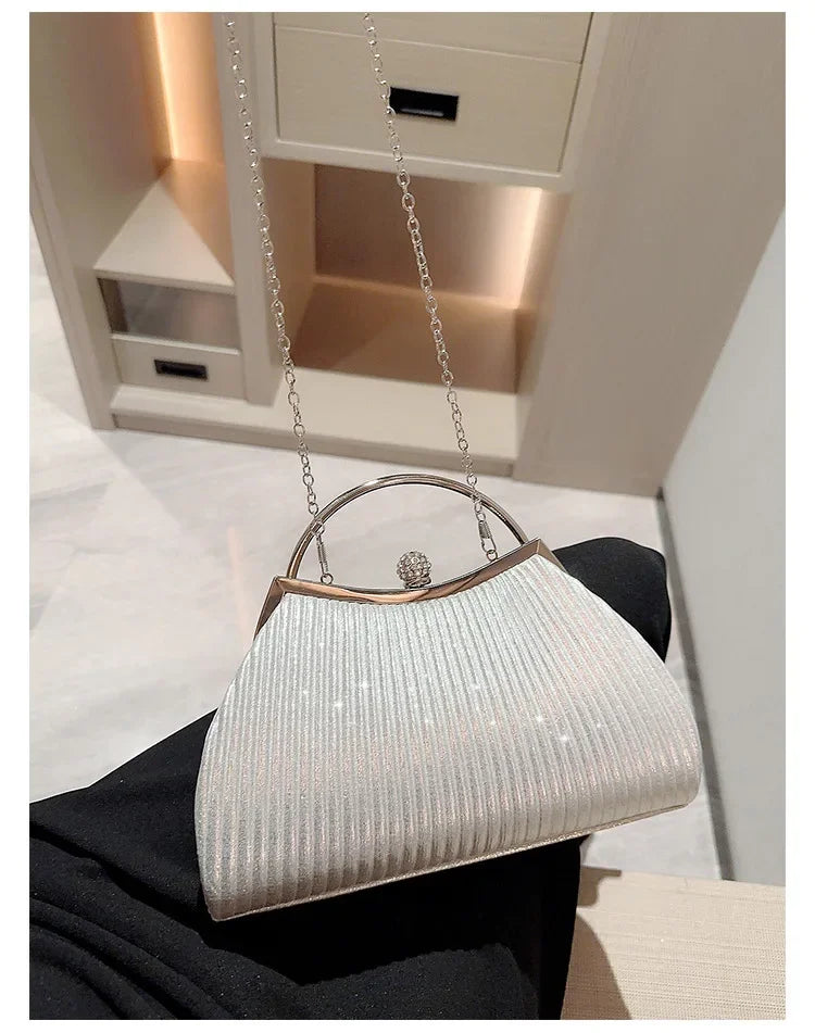 A light silver pleated glitter evening handbag with a top handle and chain strap is displayed on a black fabric.