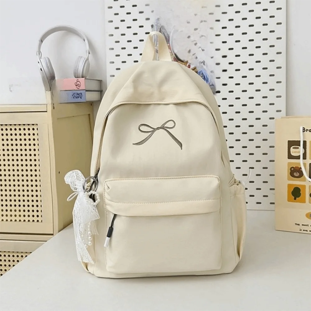 A cream-colored nylon backpack with a bow detail is displayed on a white surface next to a light-colored storage box.