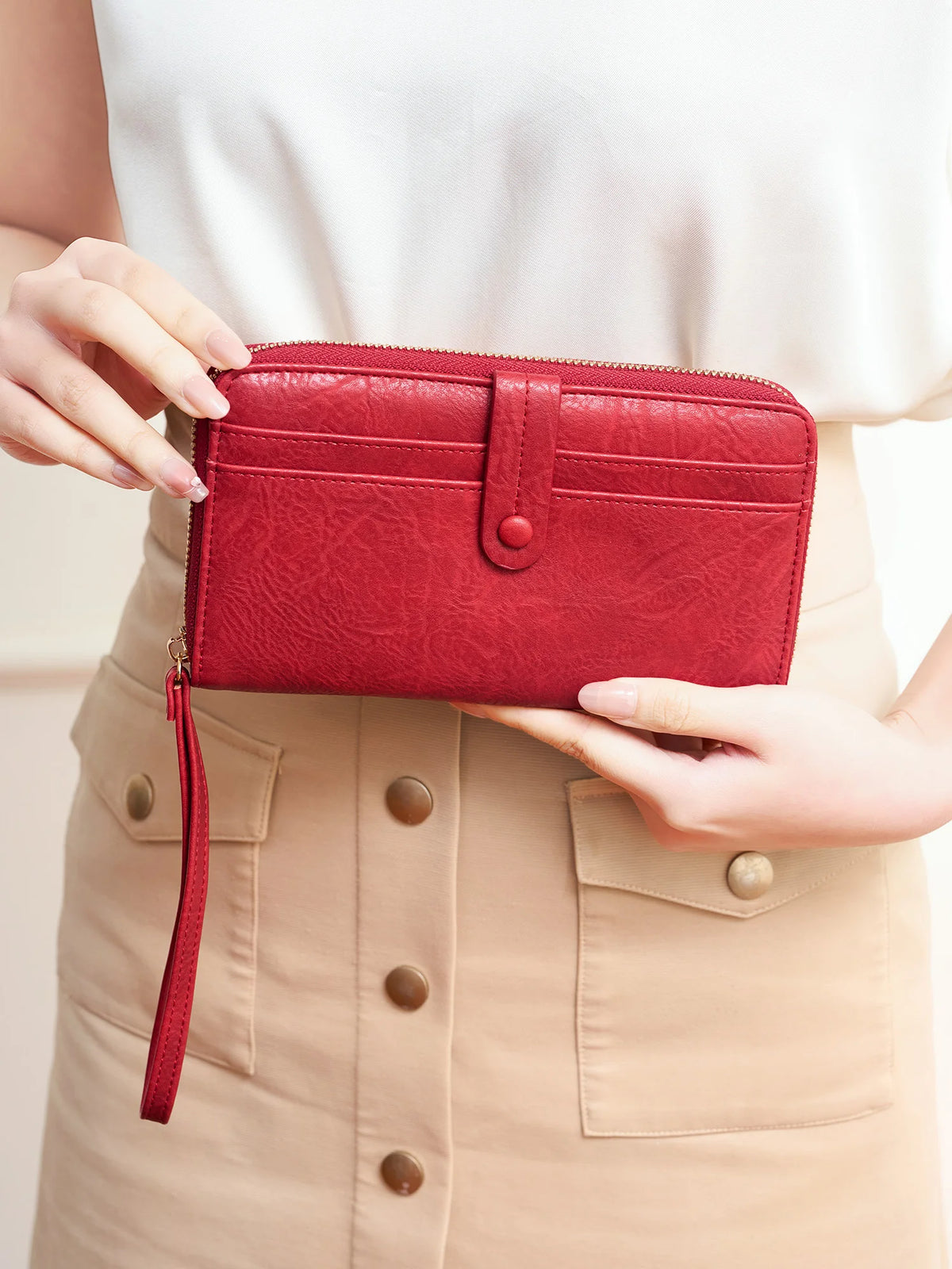 A woman holds a red PU leather zipper clutch wallet against a beige skirt.