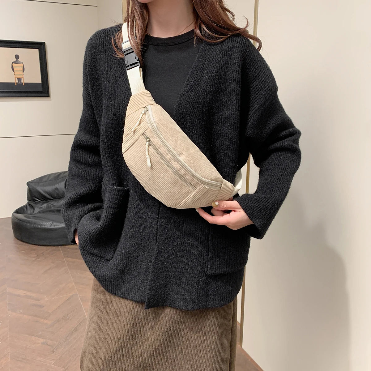 A woman is holding a beige corduroy crossbody waist bag with a white strap against a light background.