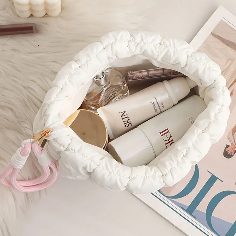 A white quilted puffer makeup bag filled with various cosmetic bottles and a pink strap, resting on a light surface.