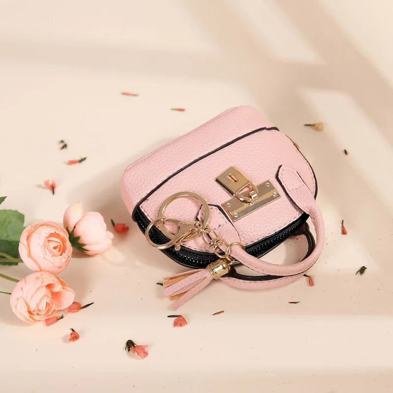 A pink portable earbud case with a gold keychain charm lies on a light surface surrounded by dried flower petals.